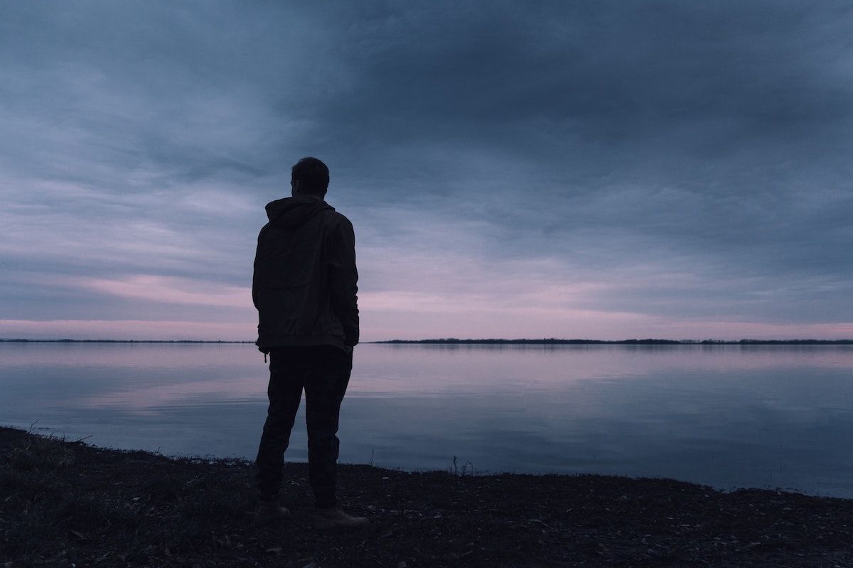 Man stood by a lake of water at dusk, staring into the distance.