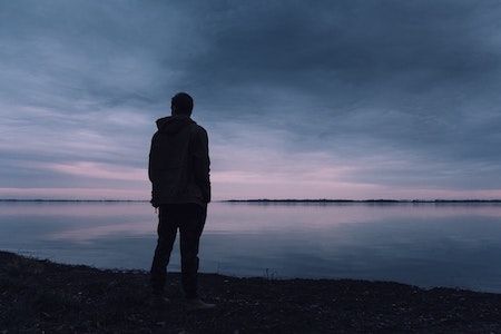 Man stood by a lake of water at dusk, staring into the distance.