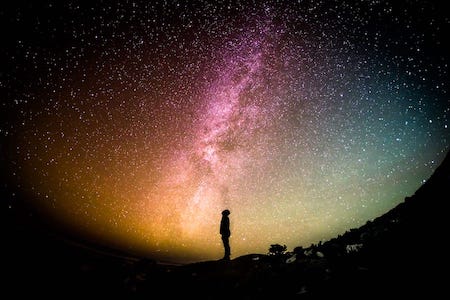 Silohuette of a young man looking up at the night sky; a colourful cosmic cloud with many stars.